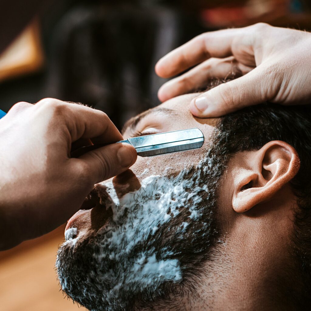 Primer plano de un barbero afeitando a un hombre con barba y crema de afeitar en el rostro